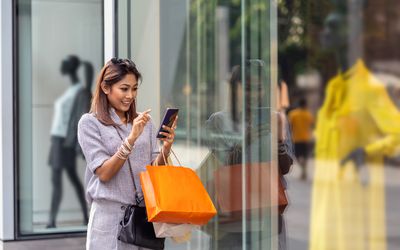 Asian woman using the smart mobile phone for check online shopping order is completed with clothes beside the glassess in store shop with happy action at department center