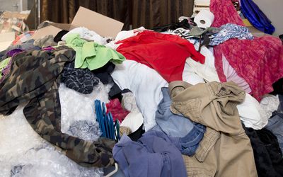 Large messy pile of household items, clothes, boxes, hangers, bubble wrap, toilet paper, pants, shirts, dresses with curtains and mirror in background