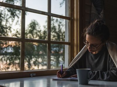 Young woman sitting at the window at a lake writing into a diary or journal