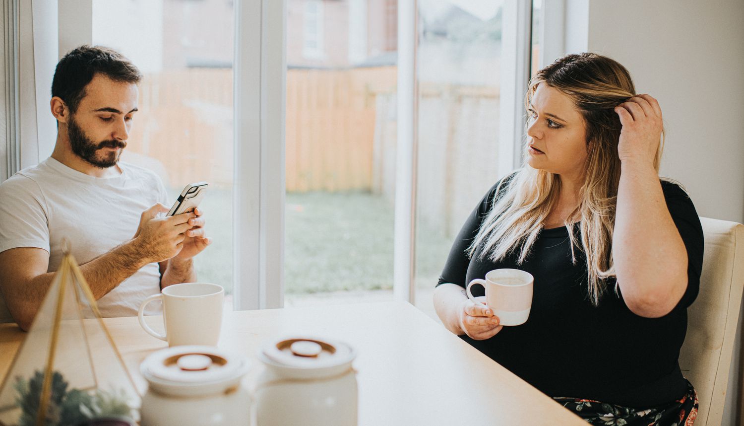 A woman and man sit at a kitchen table. The man looks down at his phone and subsequently ignores the woman. The woman looks insecure and hurt as she looks at him, hoping for some attention. Space for copy.