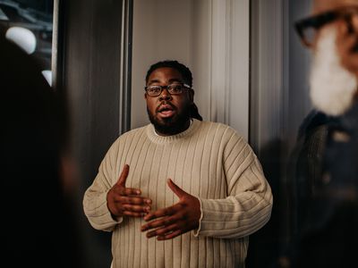 A man has a serious expression as he talks to onlookers. He uses his hands to gesture as he explains and discusses a matter with his coworkers / audience.