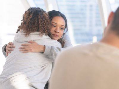 Two (2) women embrace in a comforting hug.