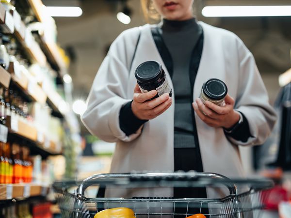 Young Asian woman with shopping cart shopping for cooking sauce in supermarket, reading and comparing the product information of two bottles of cooking sauces. 