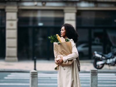 Woman with curly hair carrying a paper grocery bag crossing a street in an urban setting. She portrays a casual, everyday moment.