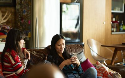 Two women relaxing at home, one texting on phone, young woman sitting beside her looking and waiting