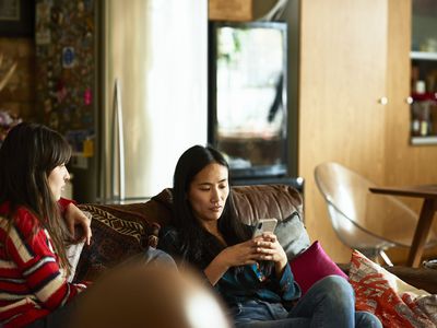 Two women relaxing at home, one texting on phone, young woman sitting beside her looking and waiting