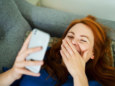 Woman lying on sofa using smart phone, smiling behind hand