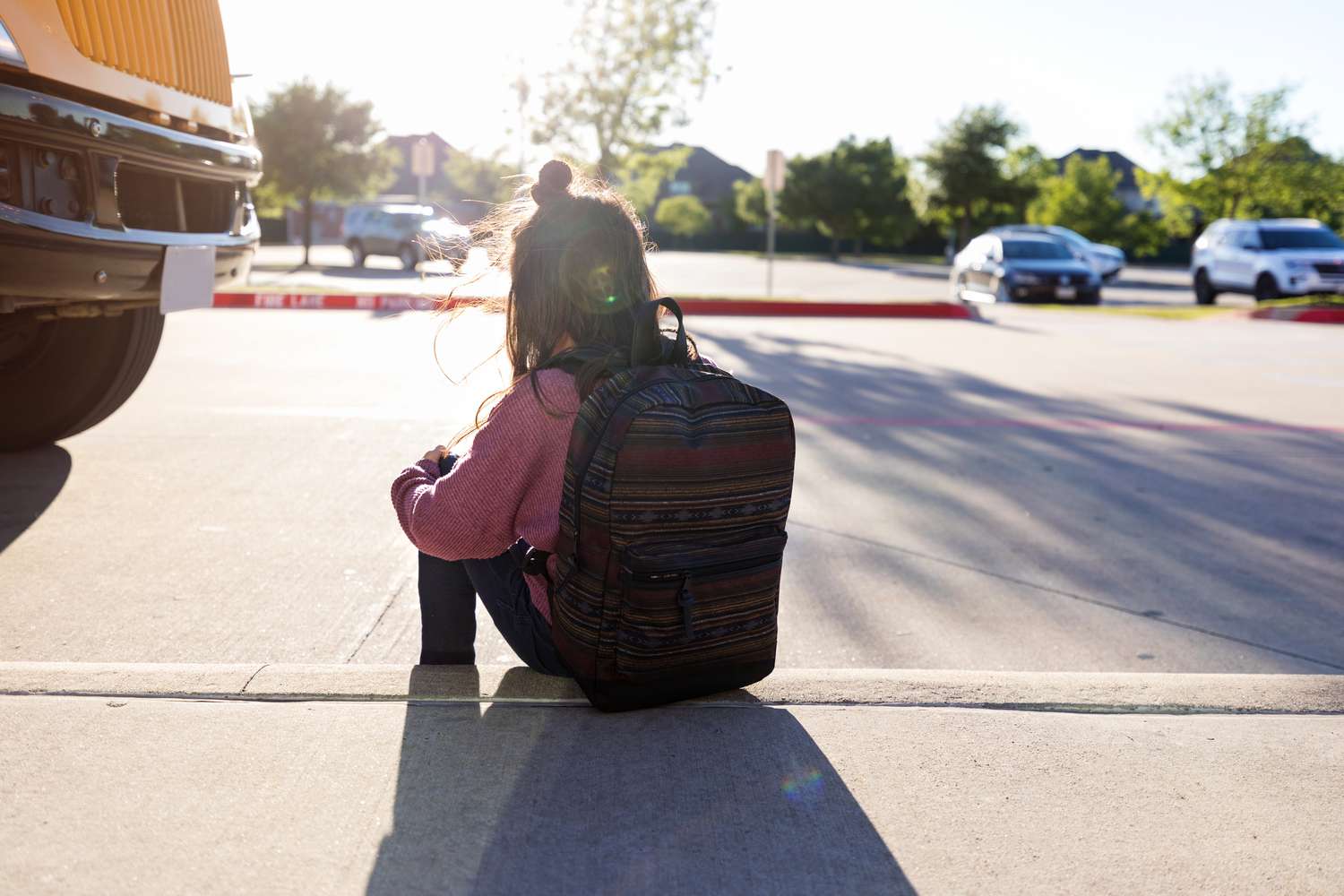 schoolgirl with a backpack hugs her knees and sits on the curb by herself at a bus stop