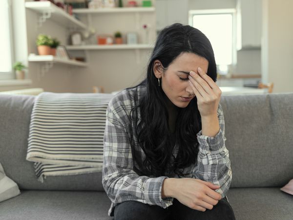 Frustrated young woman suffering from the headache while sitting on the sofa at home with an expression of being unwell, with eyes closed.