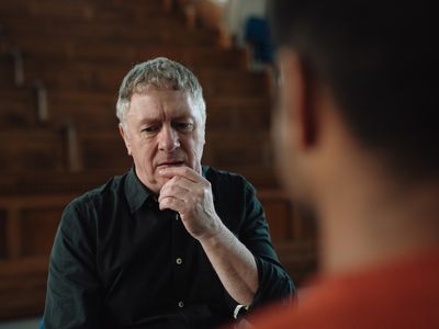 Man with grey hair and black shirt holding hand at mouth whilst thinking and talking to male therapist. Over shoulder shot