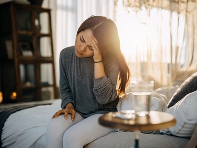 Young Asian woman sitting on the bed feeling sick and suffering from headache, a glass of water and medicine on the side table