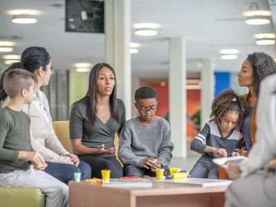 Women and kids sit around a table in a colourful facility and discuss.