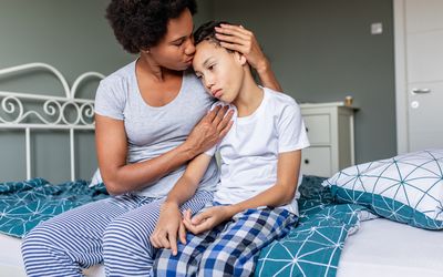 Sad boy is sitting on bed and his mother is hugging and him