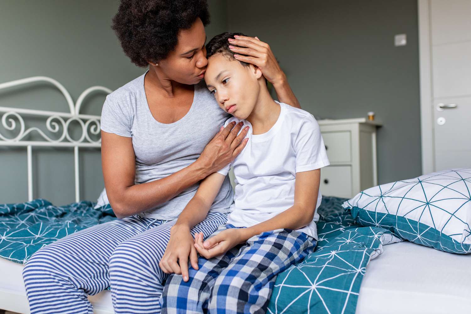 Sad boy is sitting on bed and his mother is hugging and him