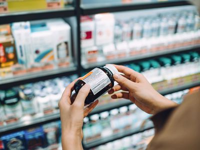 Over the shoulder view of young Asian woman browsing through medical products and reading the label on a bottle of medicine in front of the shelves in a pharmacy