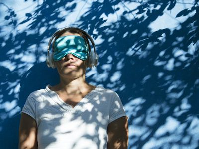 Woman in blue sleep mask and headphones listens to relaxing music for sleep against a blue background with leaf shadows. copy space