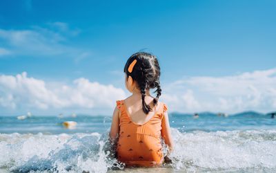 Rear view of lovely little Asian girl sitting by the seashore at the beach and being splashed by waves. Having fun at beach on a sunny Summer day