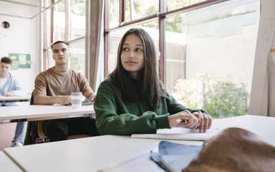 A high school student sitting at her desk by a large window in a classroom with her school mates.