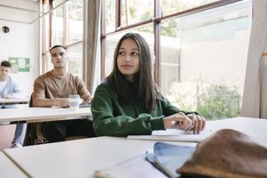 A high school student sitting at her desk by a large window in a classroom with her school mates.