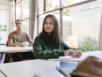 A high school student sitting at her desk by a large window in a classroom with her school mates.