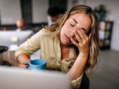 Tired and exhausted young female entrepreneur drinking coffee and rubbing eyes while feeling stressed and worried in coworking space