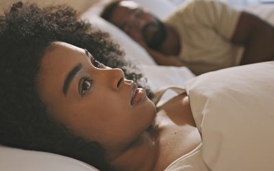 Worried female laying in bed with her husband looking anxious and concerned while thinking of her relationship issues. A man sleeping while his wife lays awake at night feeling depressed and troubled