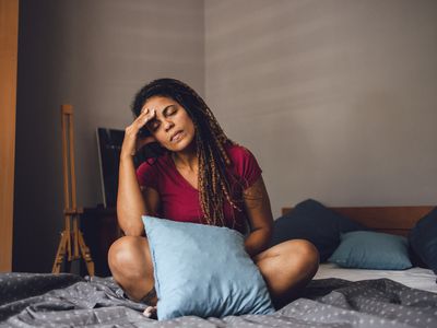 Young Latino woman feeling frustrated, sitting on a bed in lotus position with hand placed on her face.