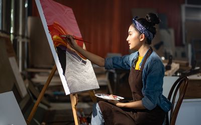 Smiling portrait on young asia female artist,she sitting in front of her big painting on canvas.Asian woman spending weekend morning painting in studio room at home