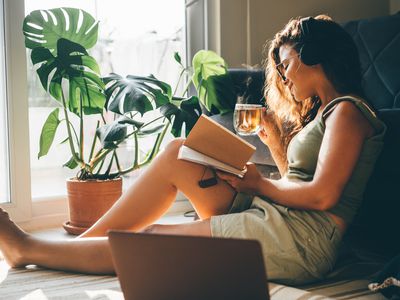 Young woman with laptop and notebook sitting in living room and drinking tea.