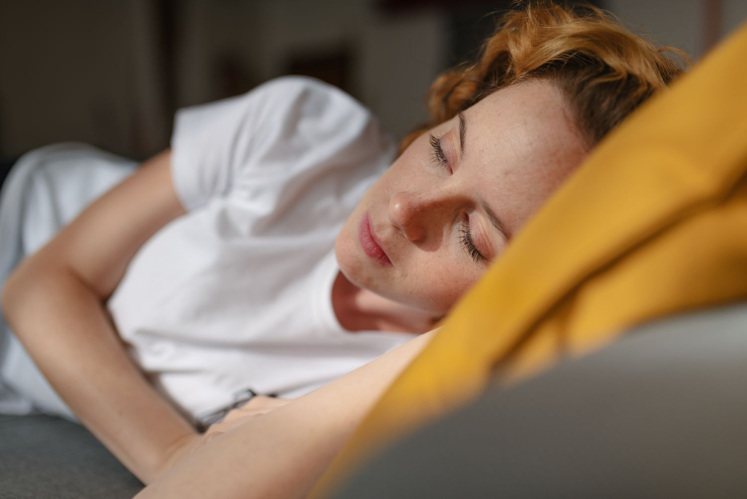 Close up view of a redhead young woman, dressed in casual clothes, lying on her side on a sofa in the living room of her house, looking away with sad face. Moment of loneliness and sadness.