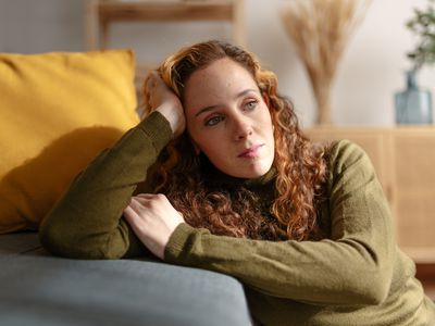 Close up view of a young woman with curly red hair sitting on the floor, leaning on a sofa and looking away with a sad face.