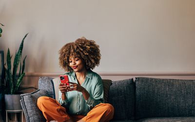 A smiling elegant African-American female using her smartphone while sitting on the cozy sofa in the living room.