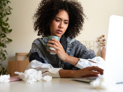 Sick African American woman working from home office using laptop drinking hot beverage covered with blanket. Sickness and working concept.