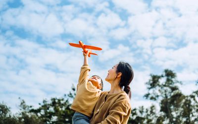 Young Asian mother and lovely little daughter spending time together outdoors, playing with airplane toy and smiling joyfully in park on a lovely sunny day against beautiful blue sky