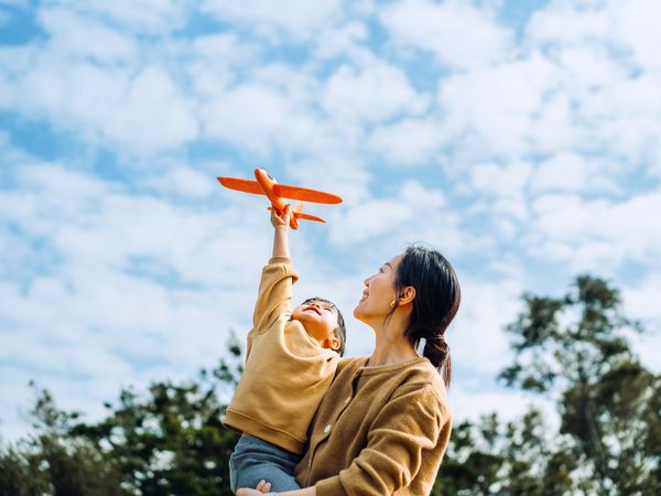 Young Asian mother and lovely little daughter spending time together outdoors, playing with airplane toy and smiling joyfully in park on a lovely sunny day against beautiful blue sky