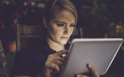 Blondhaired woman using smart device in garden, face is lit by the tablet screen.