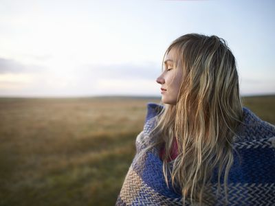 A woman stands with her eyes closed and wrapped in a blanket on a moorland.