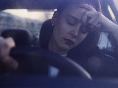 Young woman at wheel of car, eyes closed, with hand on head (grainy)