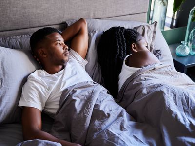 Young African man looking unhappy while lying awake next to his sleeping wife in their bed in the morning