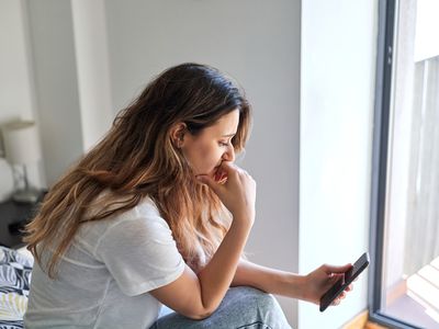 Sad and depressed woman using smart phone. She is sitting on the bed in her room. Mental health concept.