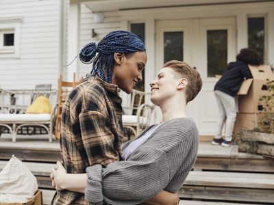 Cute queer non-binary couple embrace outside an old white house