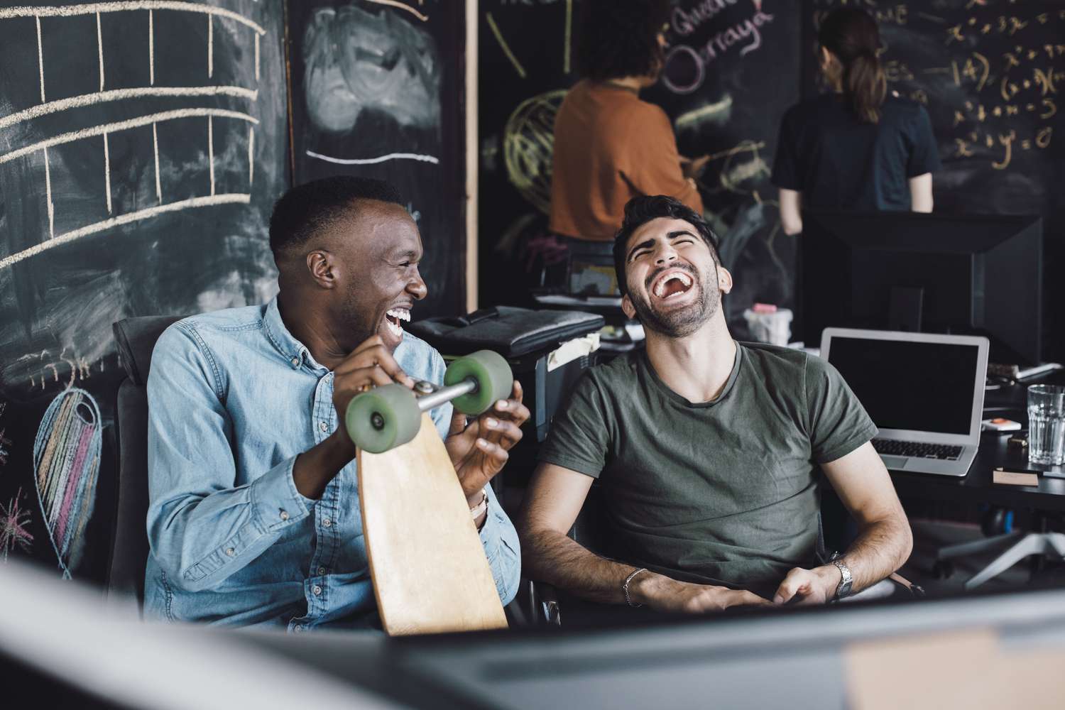 Two men sitting together laughing, one man is holding a longboard