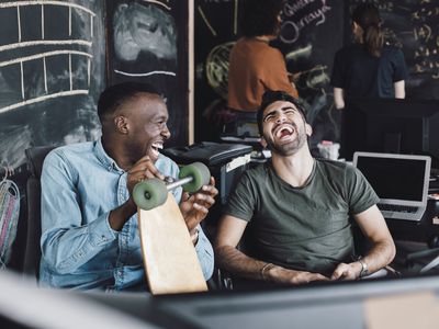 Two men sitting together laughing, one man is holding a longboard