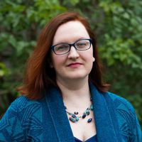 A white woman with red hair and blue glasses stands in front of green trees