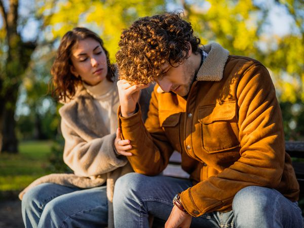 Young couple is sitting in park on sunny day. Man is sad and woman in consoling him.