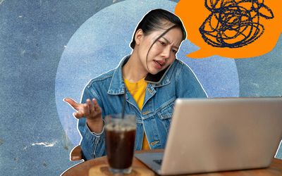 A stressed, busy, dissatisfied Asian woman in a denim jacket is on the phone with her colleague, complaining about a problem while working on her laptop remotely from a coffee shop.